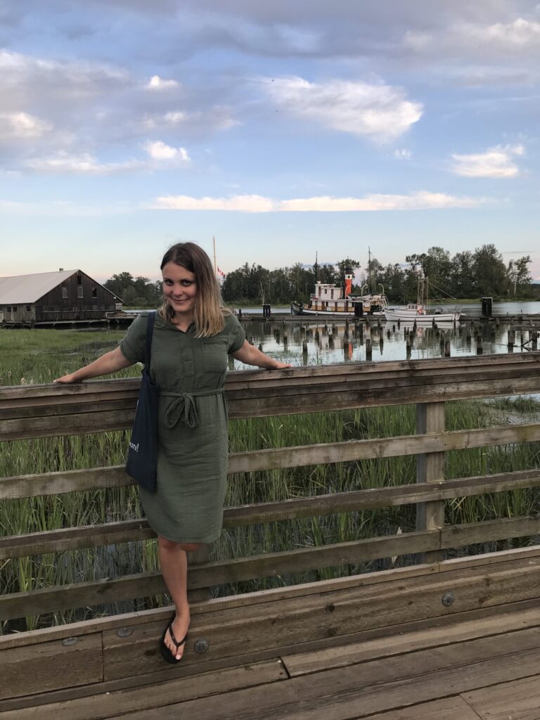 Robyn Hanson standing on the Steveston boardwalk in front of heritage boats and marshlands at Britannia Shipyards.